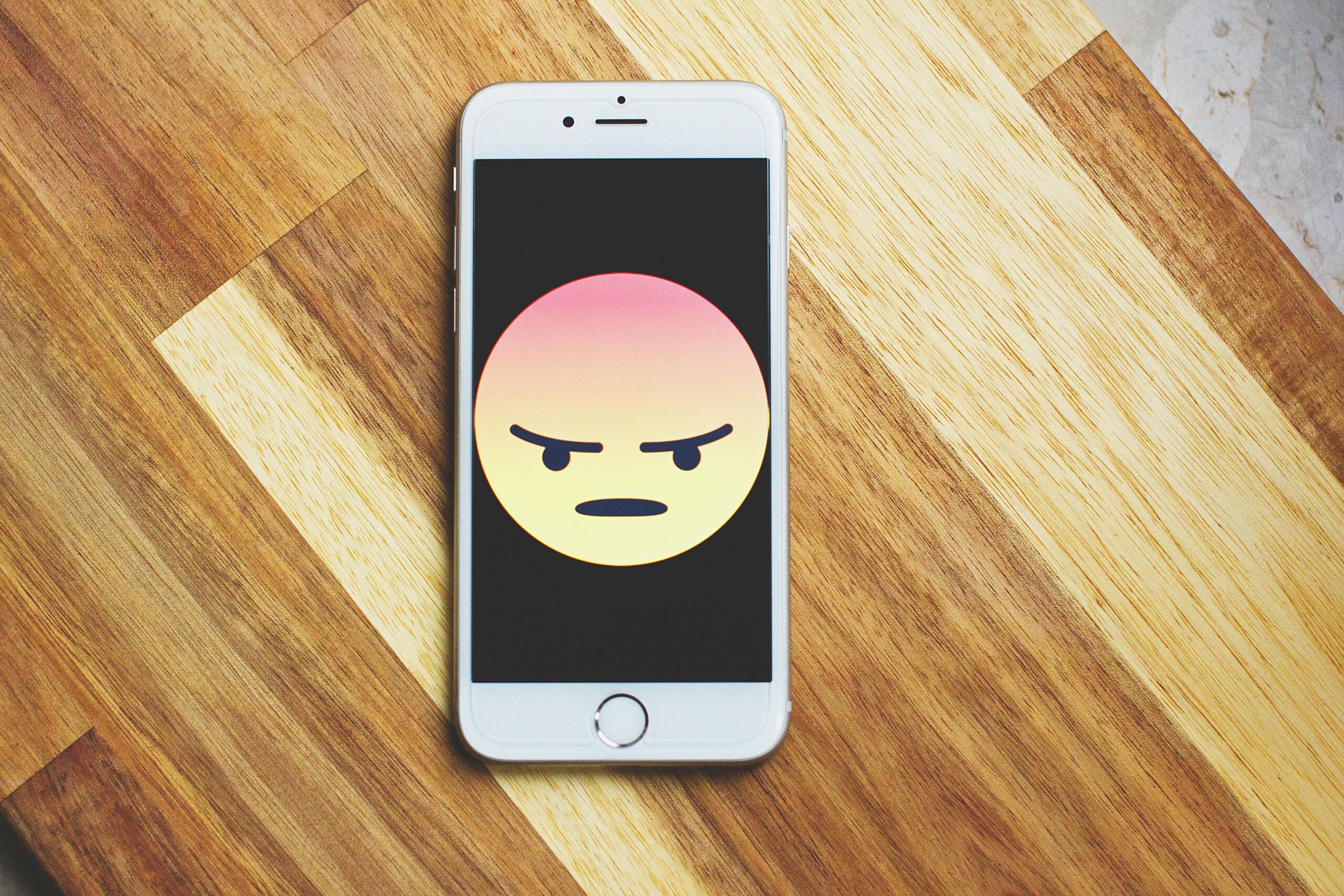 A smartphone showing an angry emoticon rests on a wooden table, emphasizing tech and emotion.